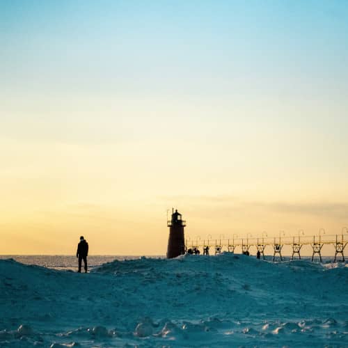 Photo avec beaucoup de neige et un homme au bord d'une falaise avec la mer, un phare et un pont dans le lointain